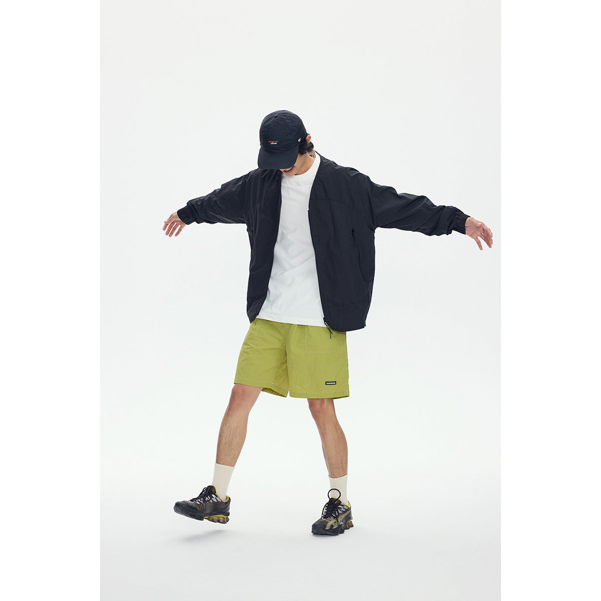 Model wears a black open jacket and lime green shorts, standing with arms outstretched.