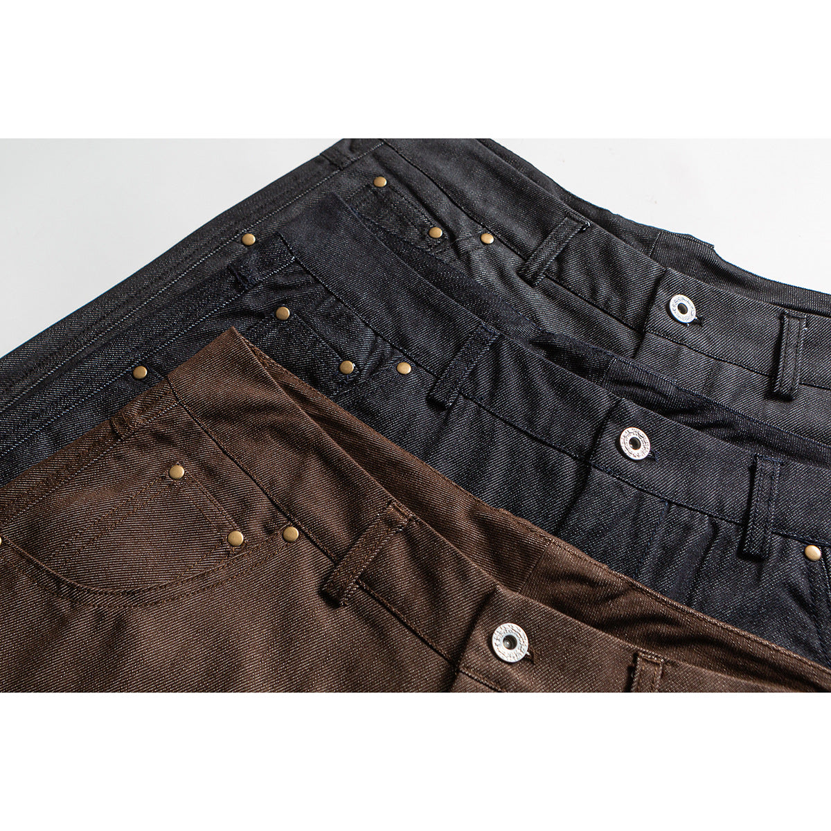 Close-up of overlapping black and brown trouser waistbands with pockets, belt loops, rivets, and metal snaps.