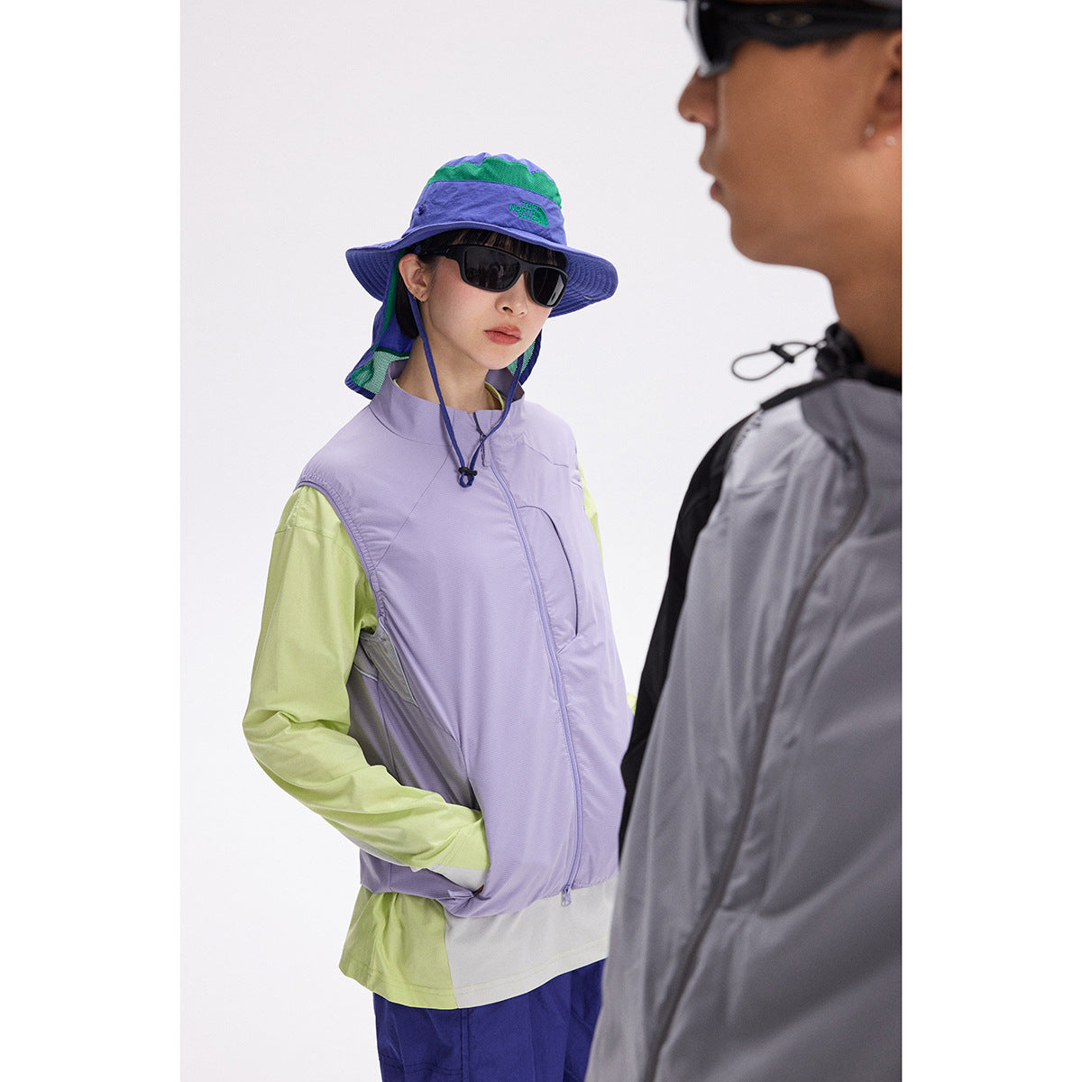 Model wearing a lavender zip vest over a lime long-sleeve shirt, with a blue-green sun hat; another person partially obscures