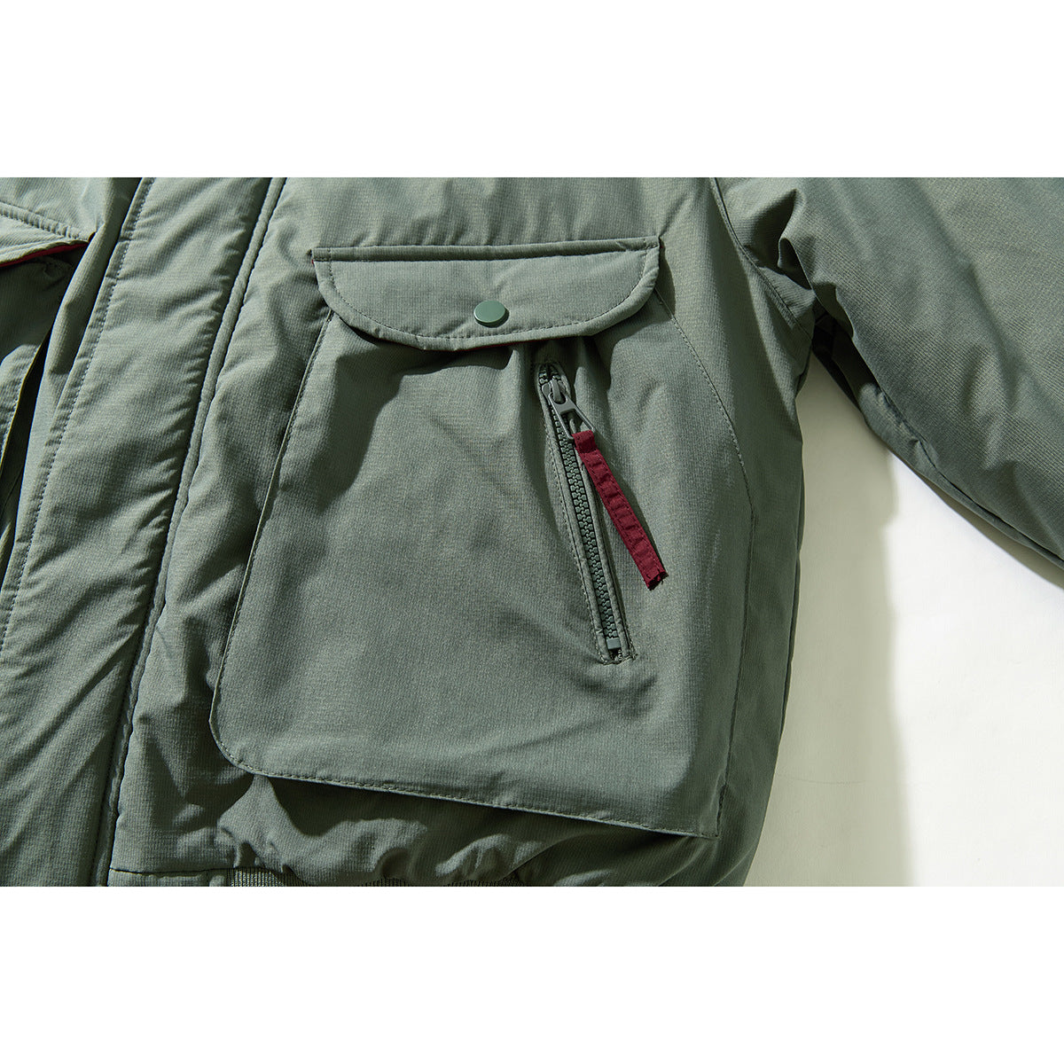 Close-up of a sage green jacket’s large flap pocket, snap button, and vertical zipper with burgundy pull.