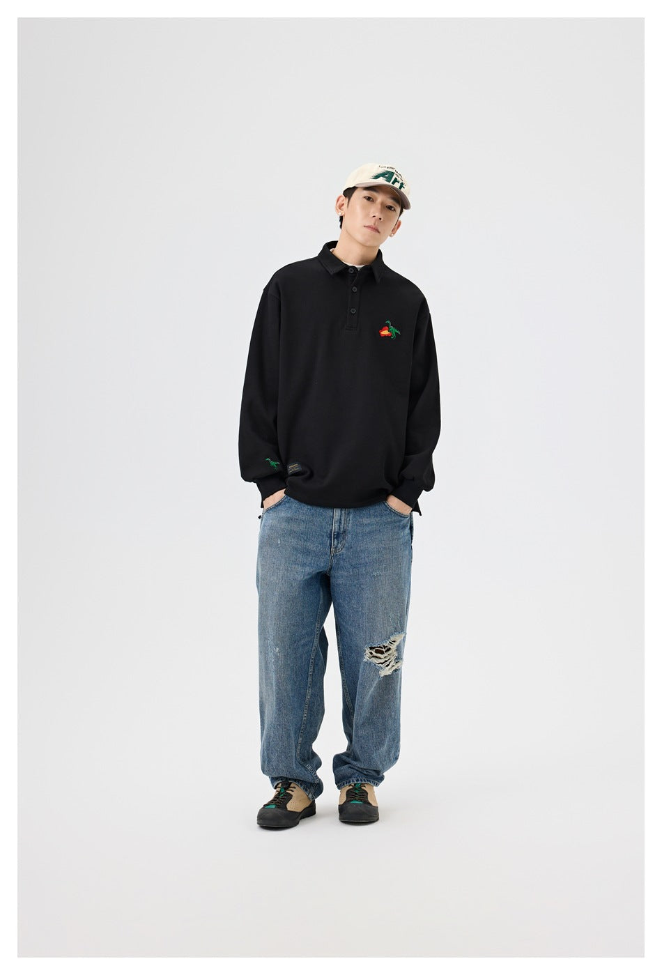 Full-length model view of a black embroidered collared pullover paired with loose distressed blue jeans and a light cap.