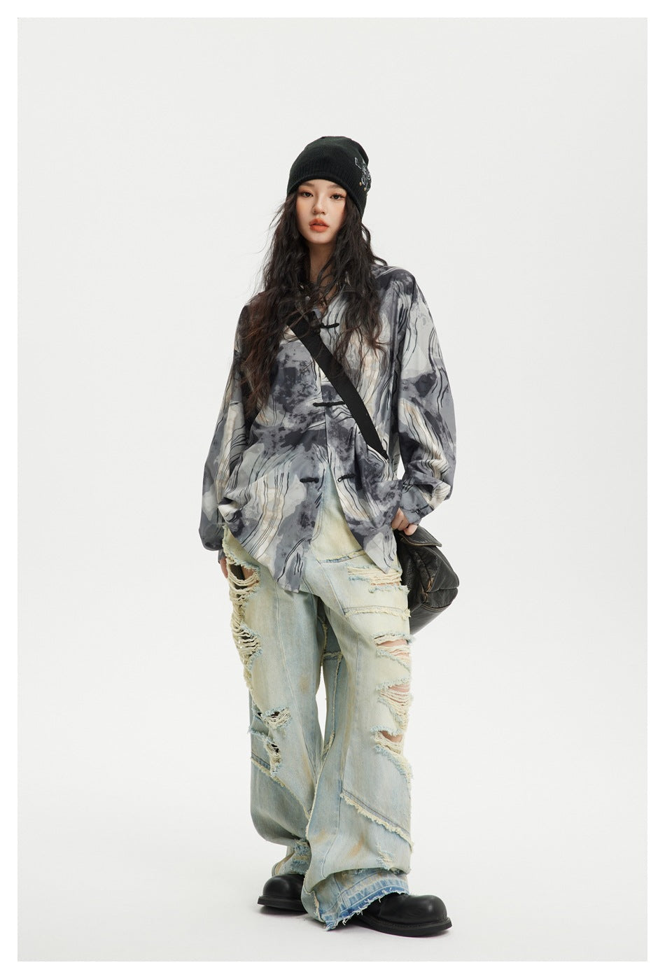 A model wears a loose gray, black, and cream abstract-pattern shirt with distressed light jeans and a black beanie.