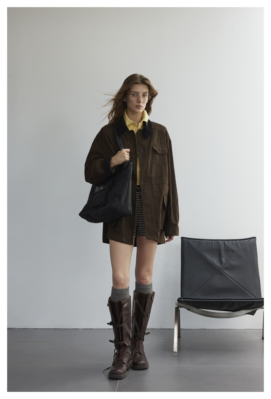 Woman wearing an oversized dark brown jacket with black collar over a yellow shirt, plaid shorts and tall brown boots
