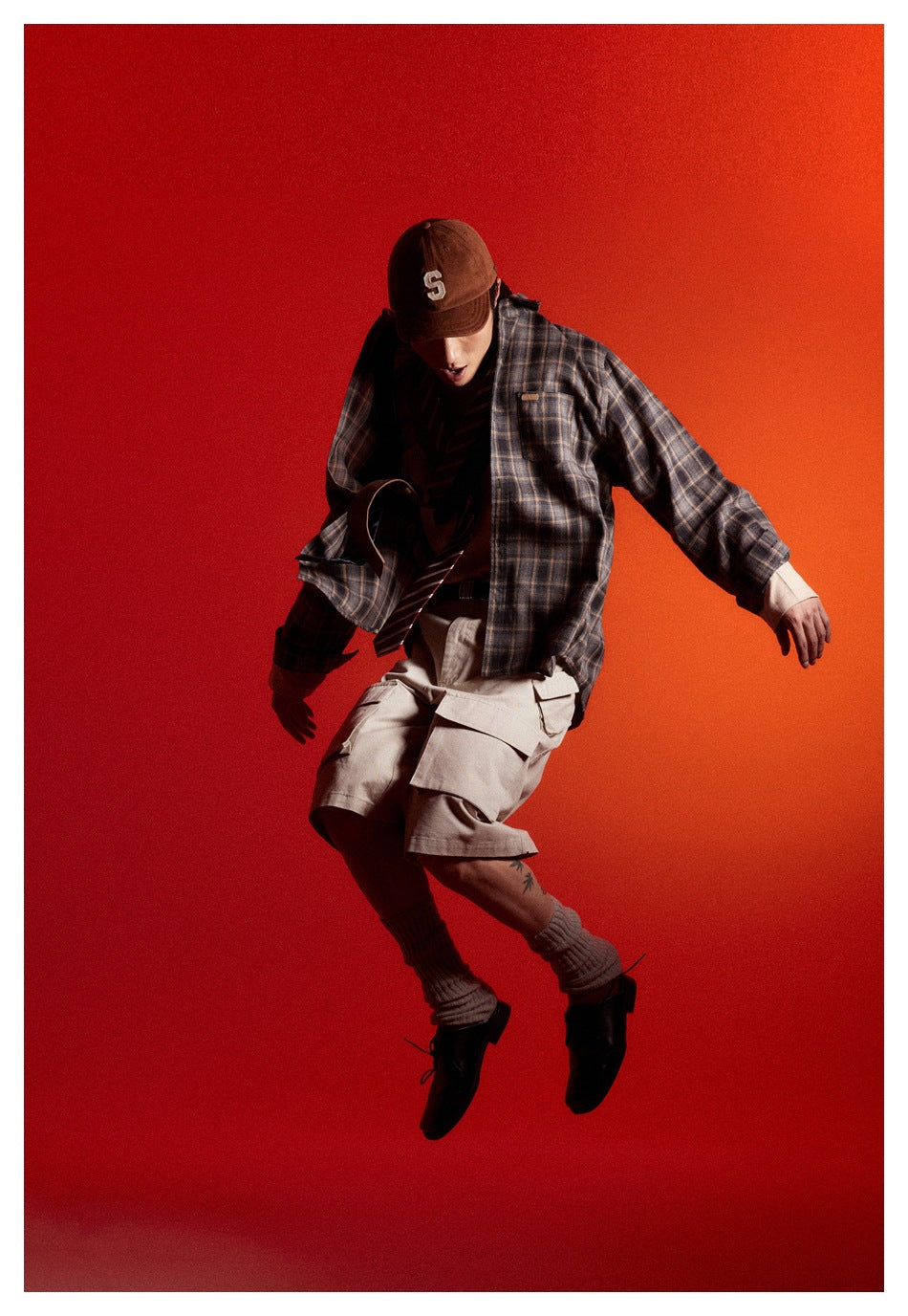 Model mid-jump in a dark brown plaid button-up shirt, beige cargo shorts, brown cap, socks and black shoes.