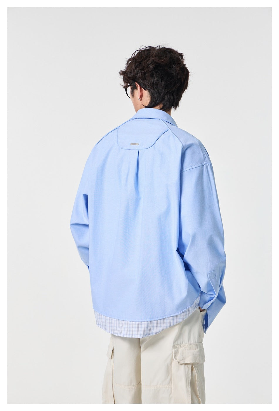 Back view of a light blue shirt on a model, showing a shoulder yoke, center pleat, and plaid hem detail.