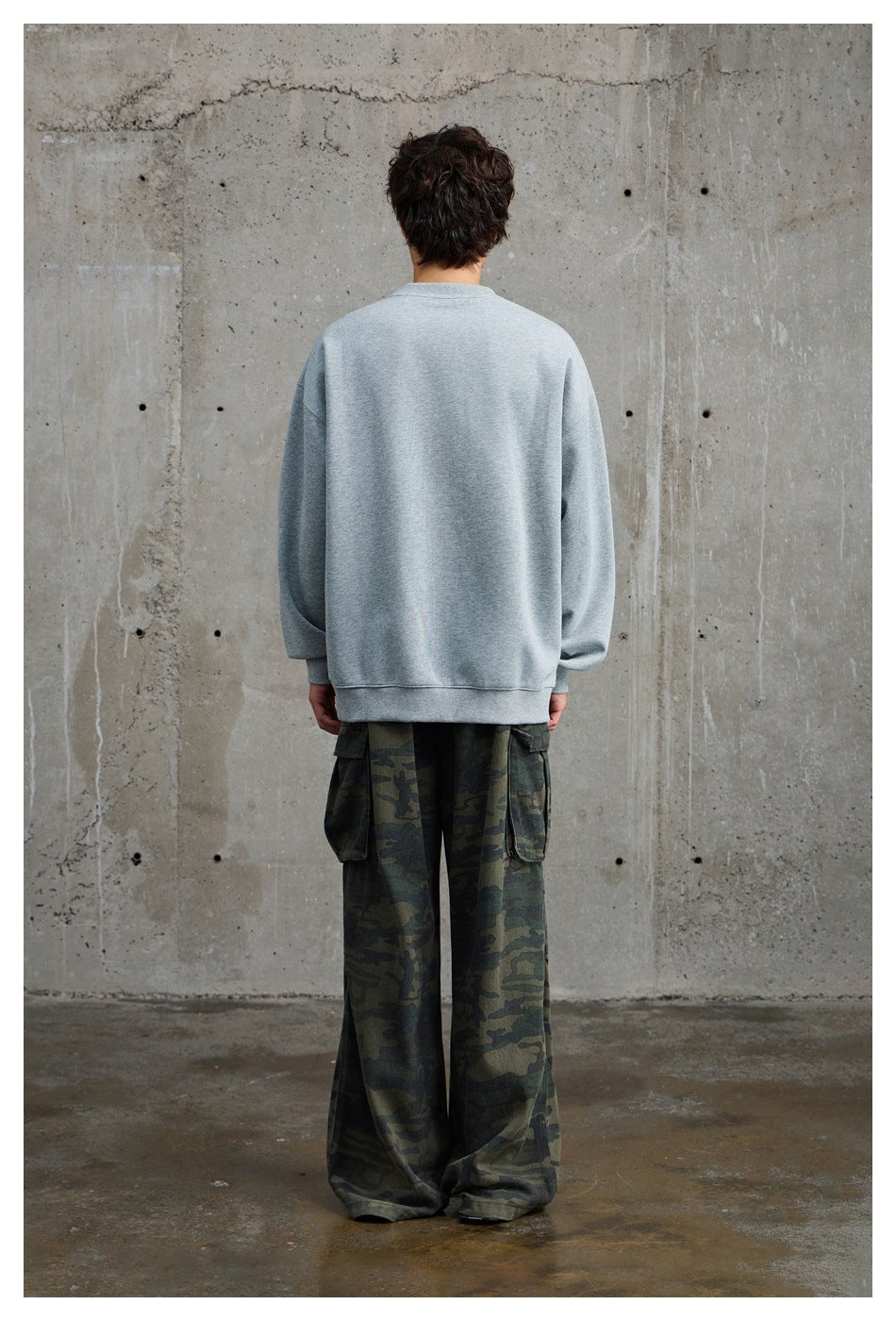 Rear view of a model wearing a plain light gray crewneck sweatshirt with oversized dark camouflage cargo pants.