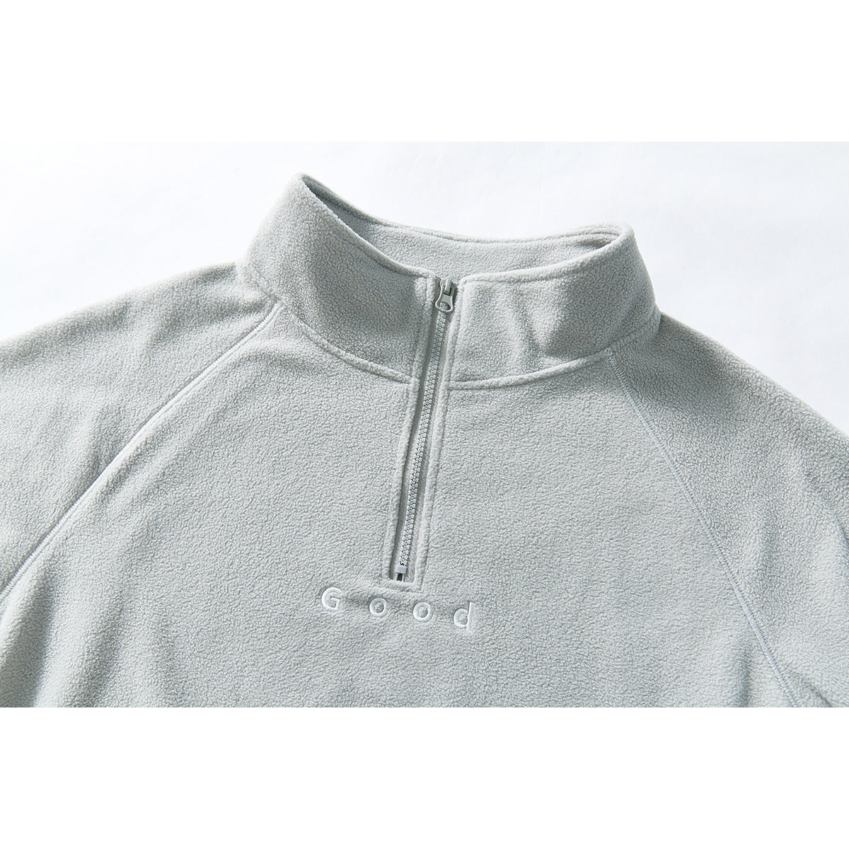Close-up of a pale gray quarter-zip pullover with a stand collar and white embroidered “Good” lettering.