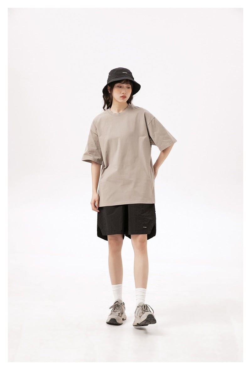 Model wearing a loose taupe T-shirt, black shorts, a black bucket hat, white socks, and gray sneakers.