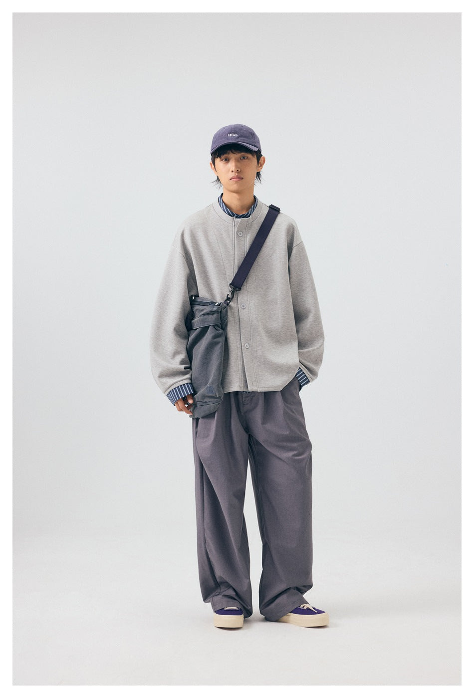 Model standing in front view wearing gray wide-leg trousers with a gray top, cap and shoulder bag