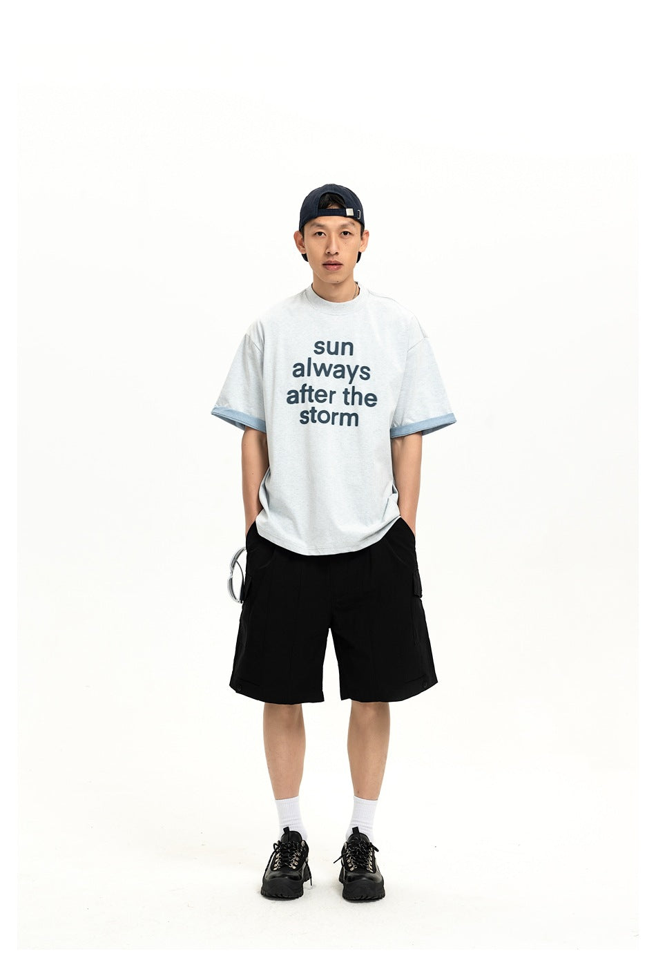 Model wearing a light gray T-shirt printed “sun always after the storm” and black cargo shorts, front view.