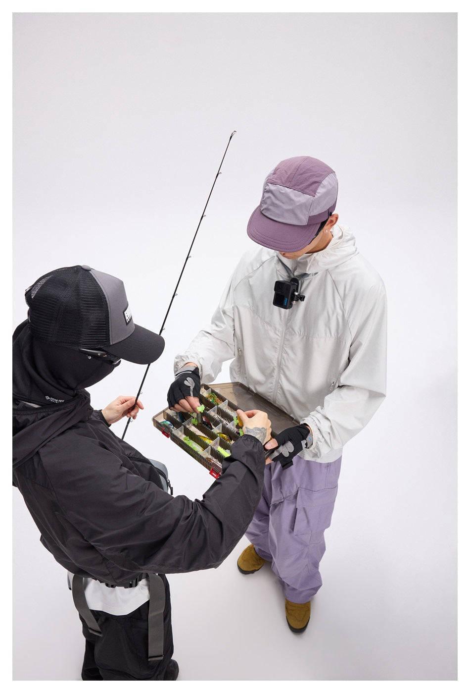 Two models inspect a tackle box; one wears a white hooded jacket and lavender cargo pants, the other a black outfit.