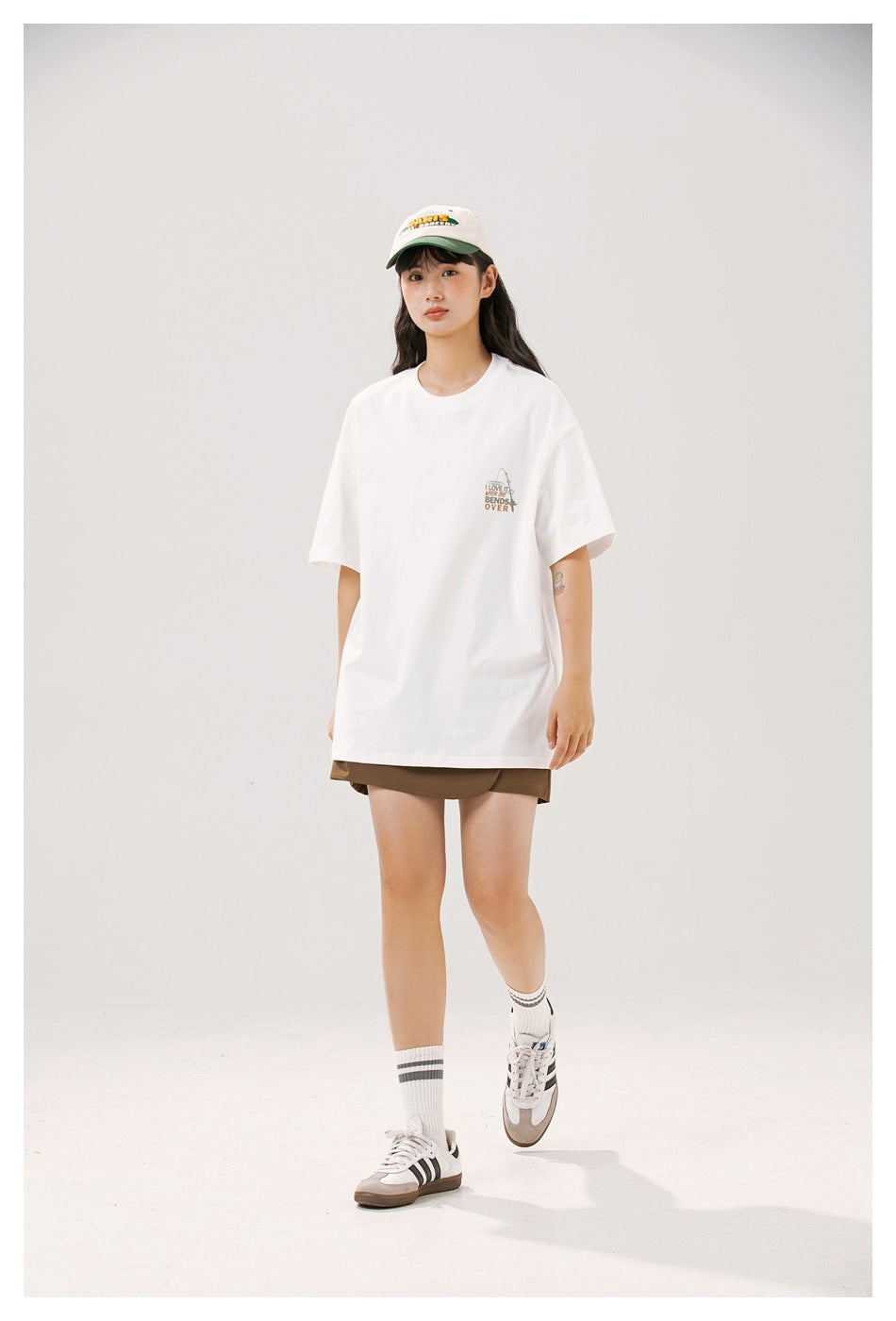 Model walking in a white relaxed-fit T-shirt with a small chest graphic, brown shorts, a cap and sneakers