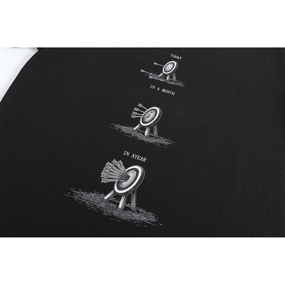 Close-up of a black T-shirt print showing targets with arrows and the English phrases “TODAY,” “IN A MONTH,” and “IN A YEAR.”