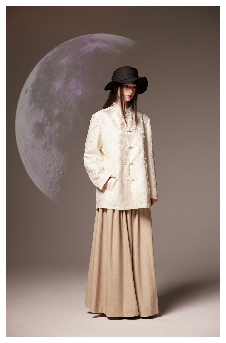 Model in an ivory frog-button jacket, beige wide-leg bottoms, and a black wide-brim hat against a moon backdrop.