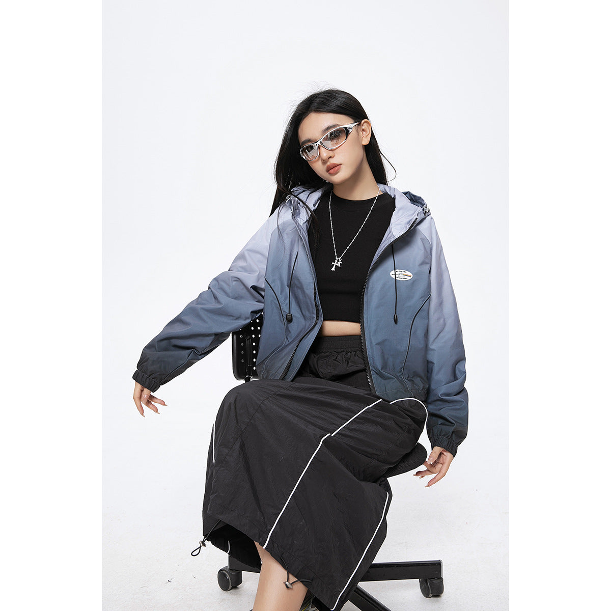 Seated model wearing a blue-gray hooded zip jacket over a black top and a long black skirt with white piping.