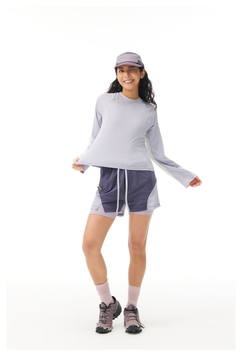 Woman wearing a lavender long-sleeve top with contrast sleeve panels, shorts and a cap, front view