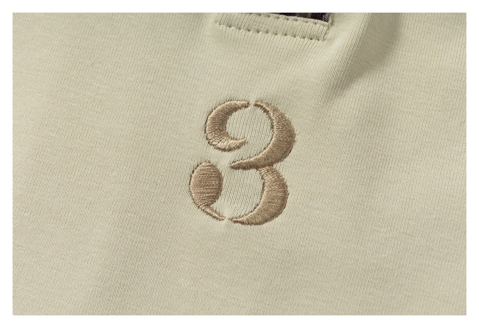 Close-up of a tan embroidered emblem on cream fabric, with a small plaid detail near the top edge.
