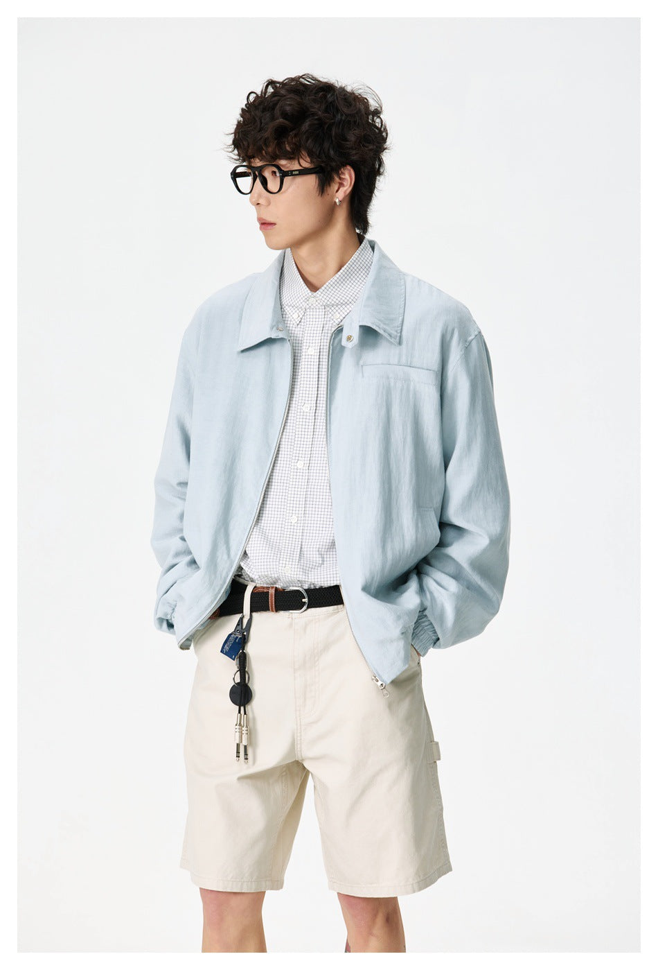 Model wearing an open pale blue jacket with a wide collar and chest pocket, paired with a checked shirt and cream shorts.