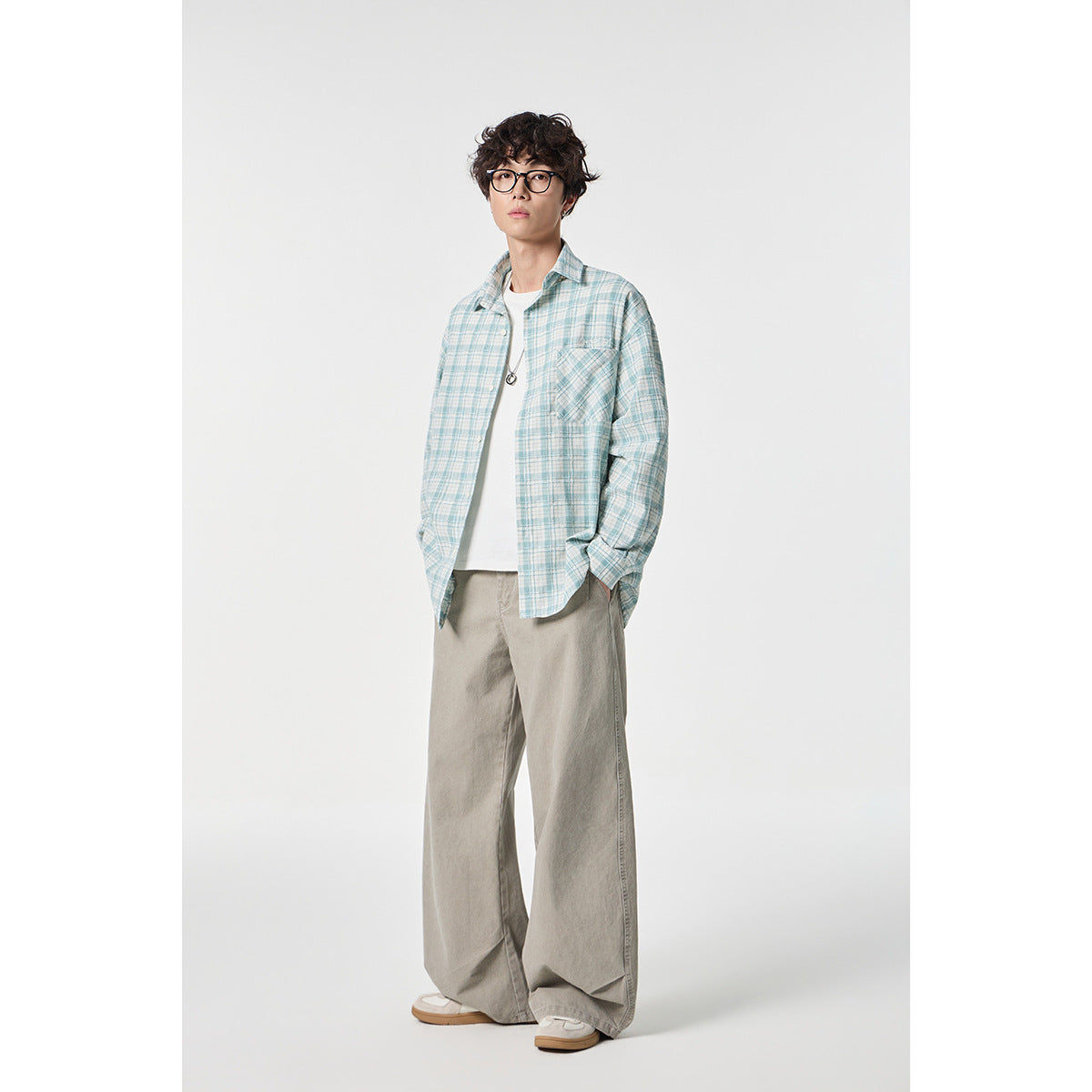 Model wearing a pale blue-and-white plaid button-up shirt over a white tee with loose taupe trousers.