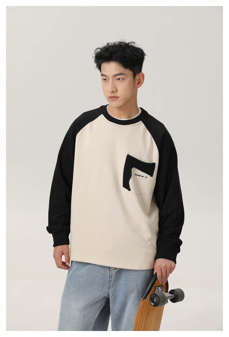 Closer front view of a cream sweatshirt with black raglan sleeves and a contrasting chest pocket, worn by a model.