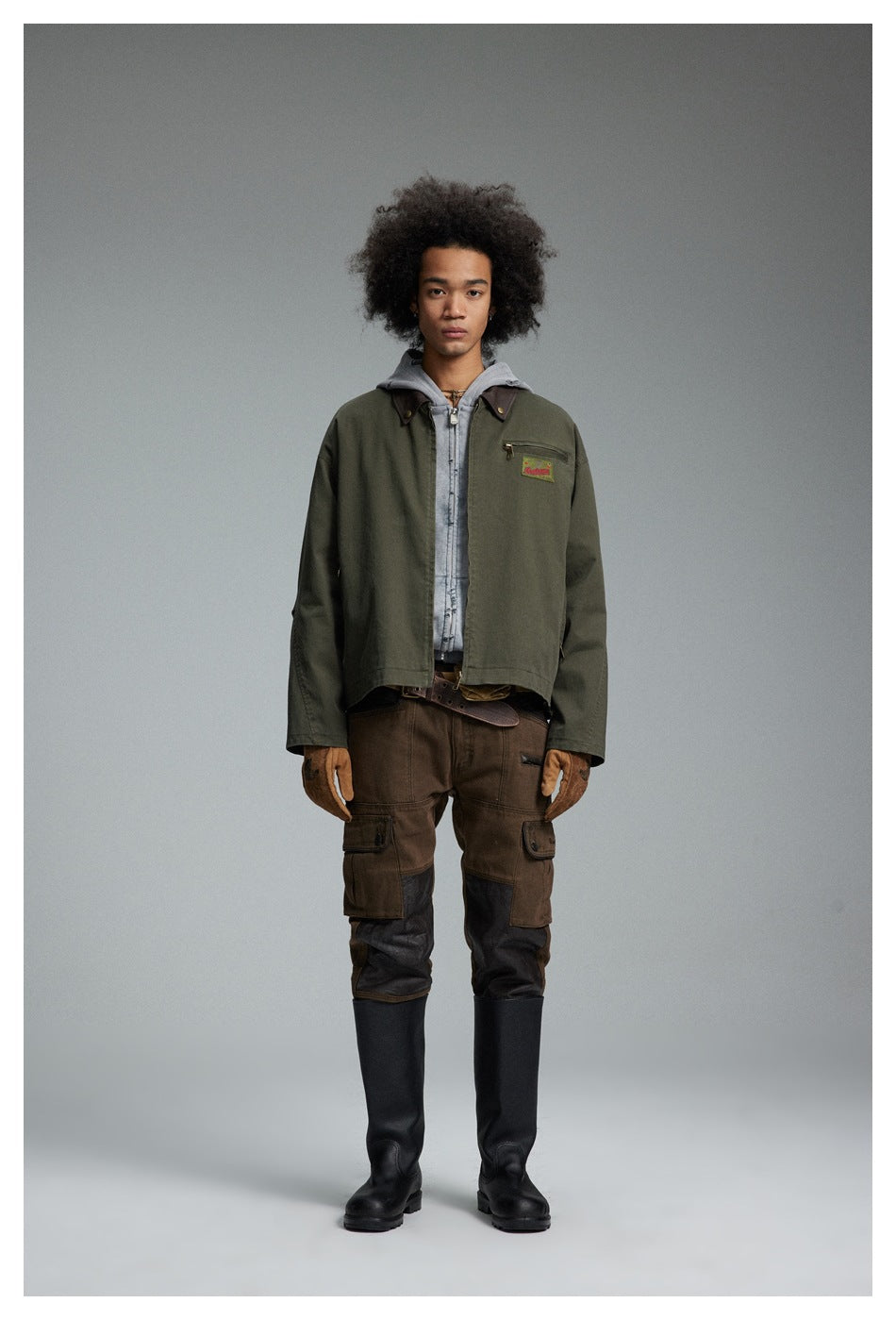 Model wearing the army green zip jacket over a gray hoodie with brown cargo pants and black tall boots, front view
