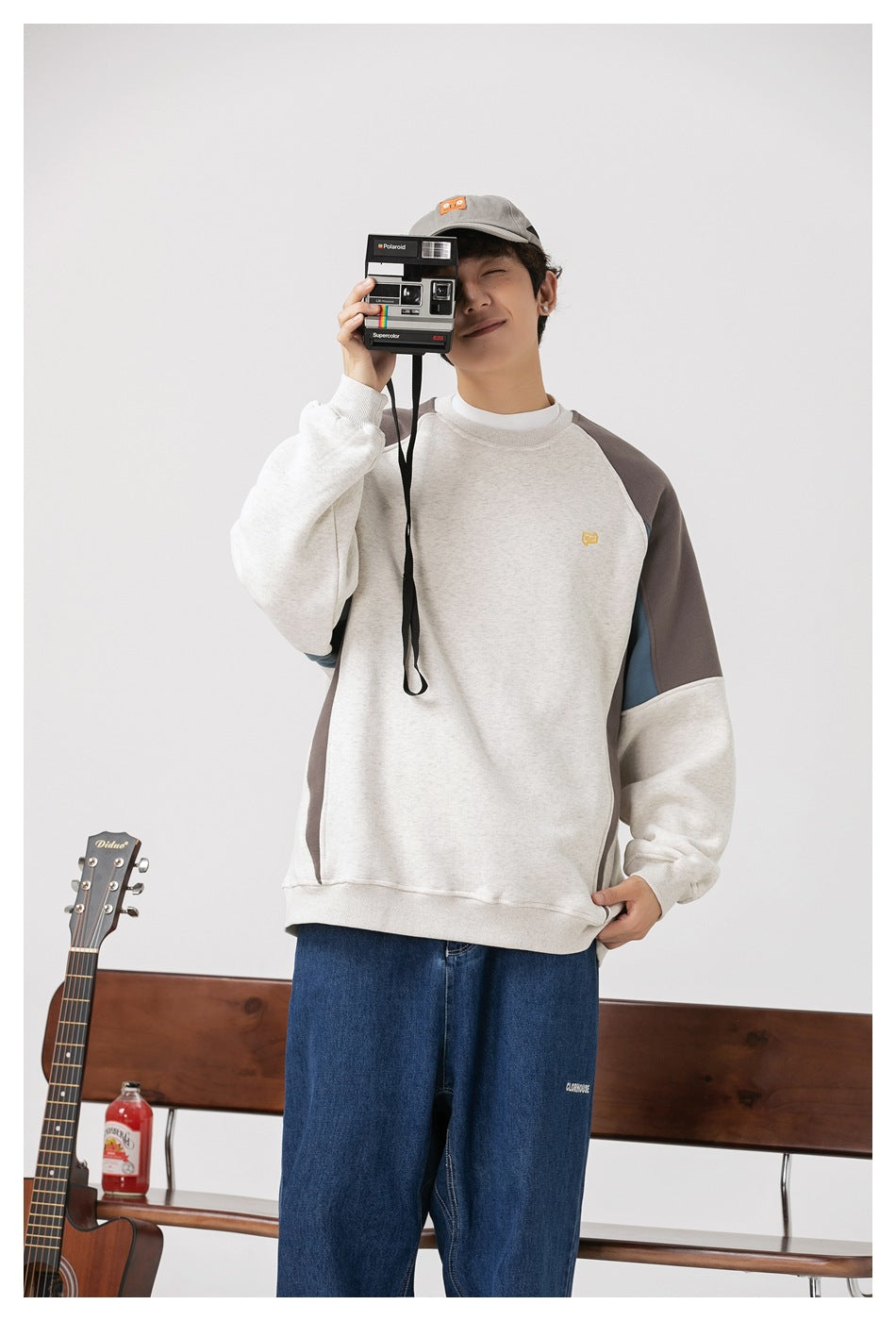 Model wearing light gray colorblock crewneck sweatshirt with jeans, holding an instant camera in front of his face