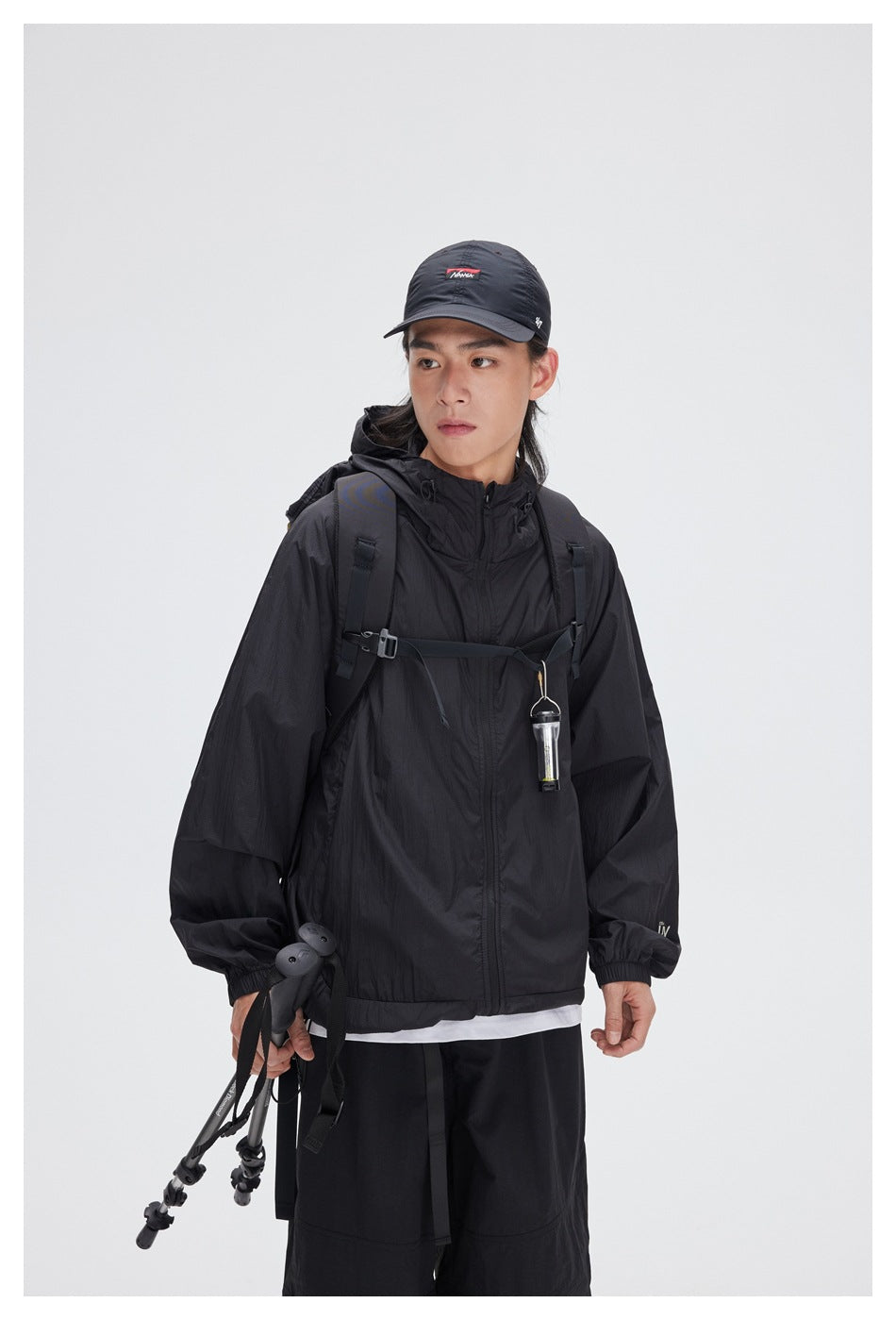Model in a black hooded jacket and cap, shown from the waist up with a backpack and trekking poles.