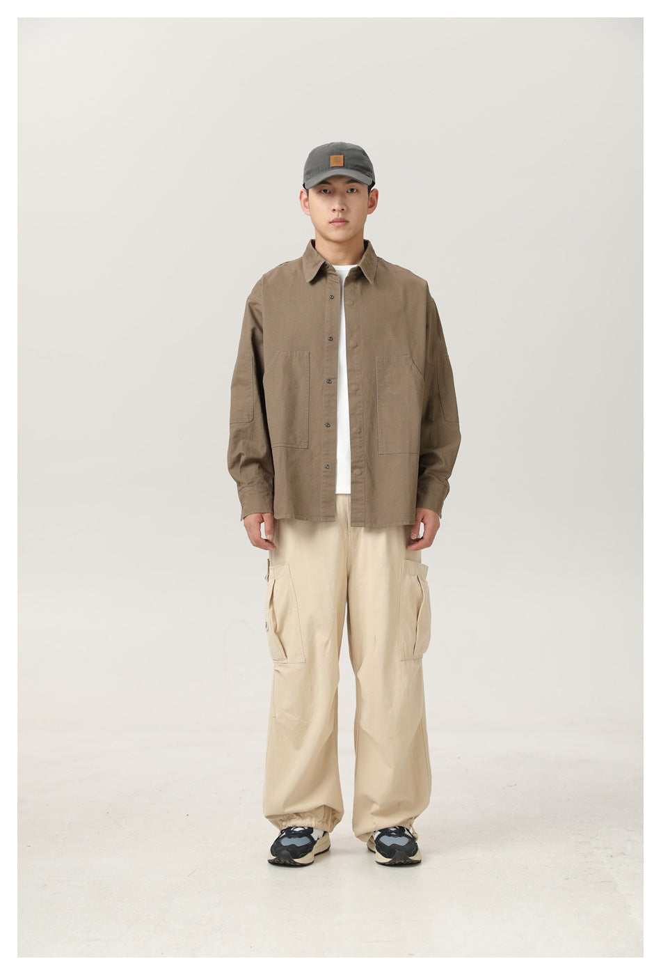 Man wearing olive-brown button-up shirt open over a white tee with beige cargo pants and a gray cap, front view