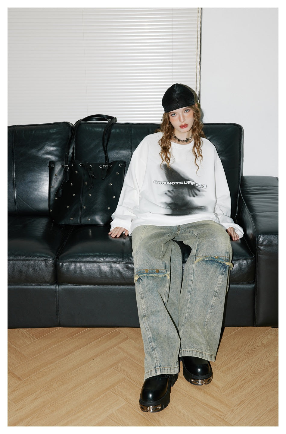 Model seated on a black sofa wearing a white graphic top and faded wide-leg jeans.