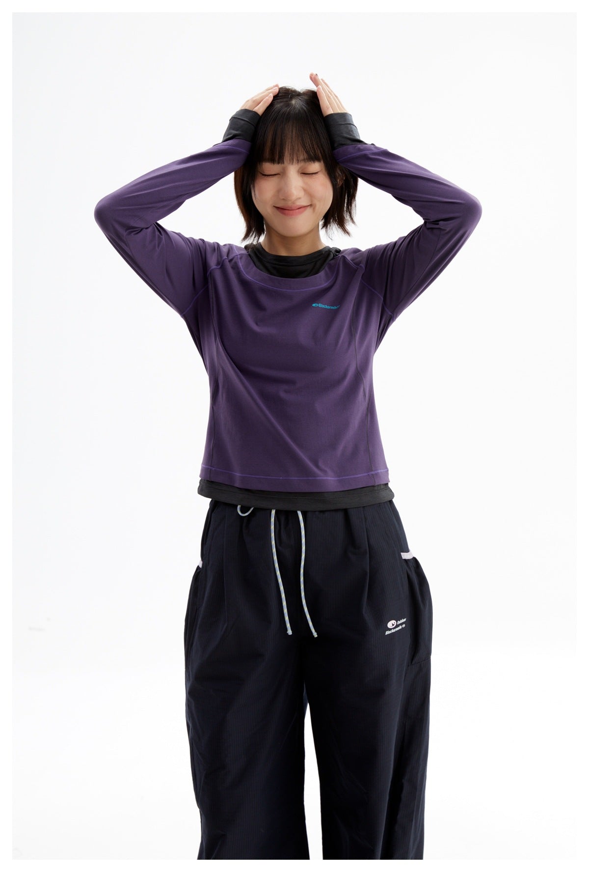 Model wearing a purple long-sleeve top layered over black and loose black pants, arms raised.