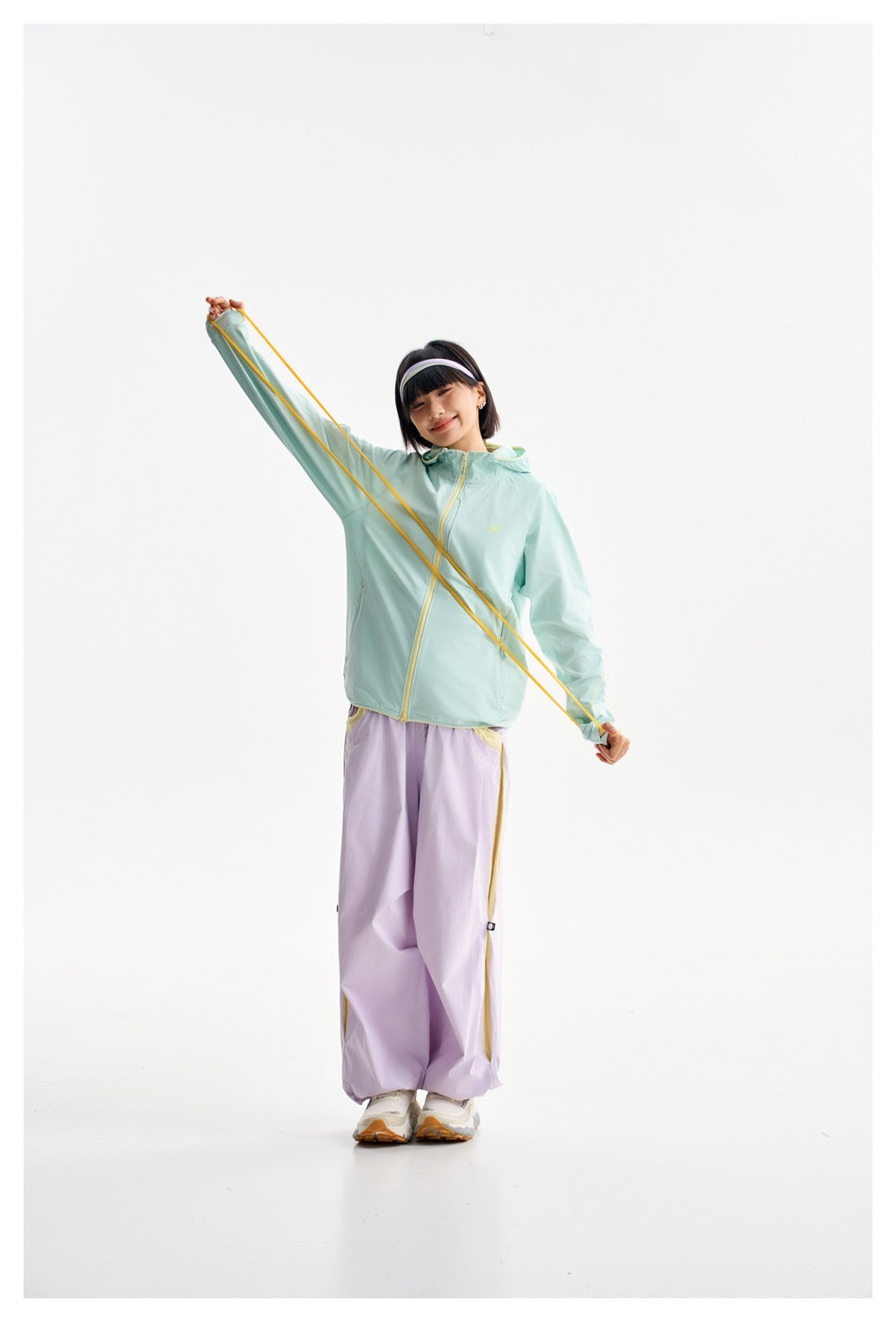 Model in a mint hooded jacket and lavender pants stretches a yellow cord diagonally across the jacket.