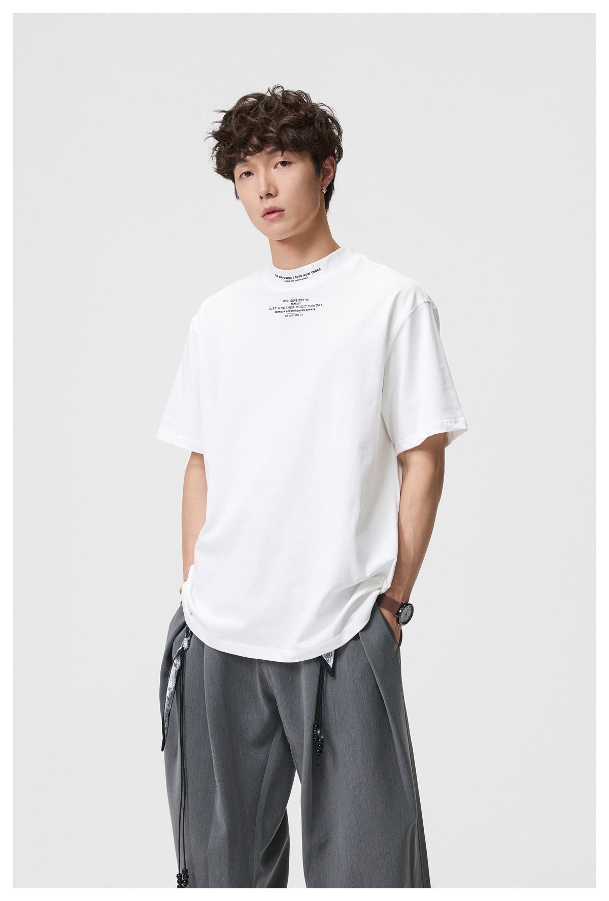 Man wearing a white mock-neck short-sleeve T-shirt with small text print, paired with gray pleated trousers
