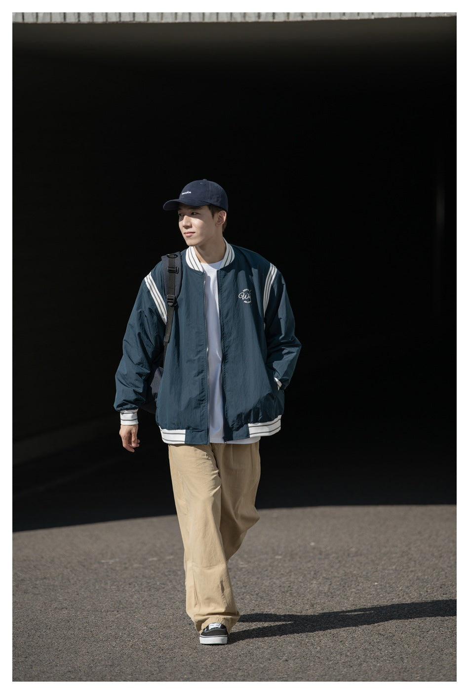 Man walking outdoors wearing a navy striped-trim bomber jacket over a white tee with khaki pants and a cap