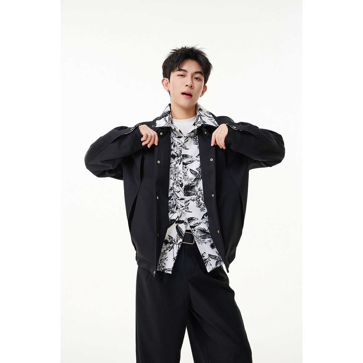 Model wearing a black snap-front jacket over a black-and-white botanical print shirt and black trousers.