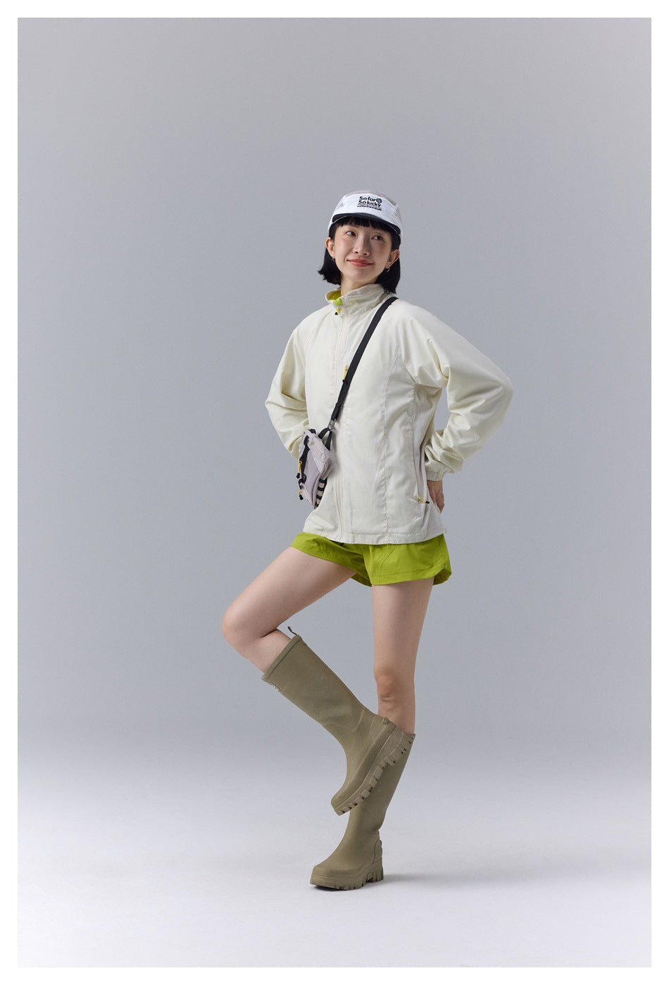 Model wearing an ivory zip-up outdoor jacket and lime shorts with a white mesh cap, crossbody bag, and tall olive boots.