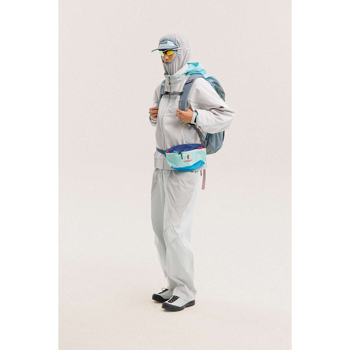 Model wearing a light gray hooded jacket and pants with a turquoise backpack and waist pouch.