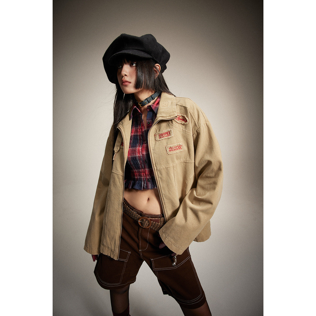 Model in a tan patch-decorated zip jacket, red plaid crop top, and brown cargo shorts, shown at an angle.