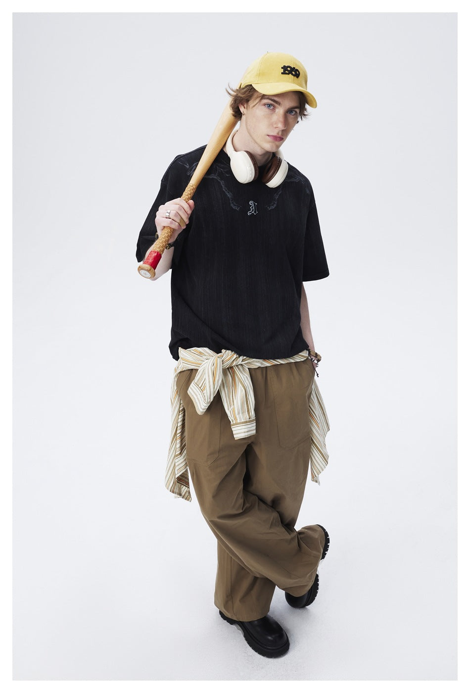 Model wearing an oversized black graphic T-shirt and olive trousers, holding a bat over one shoulder.
