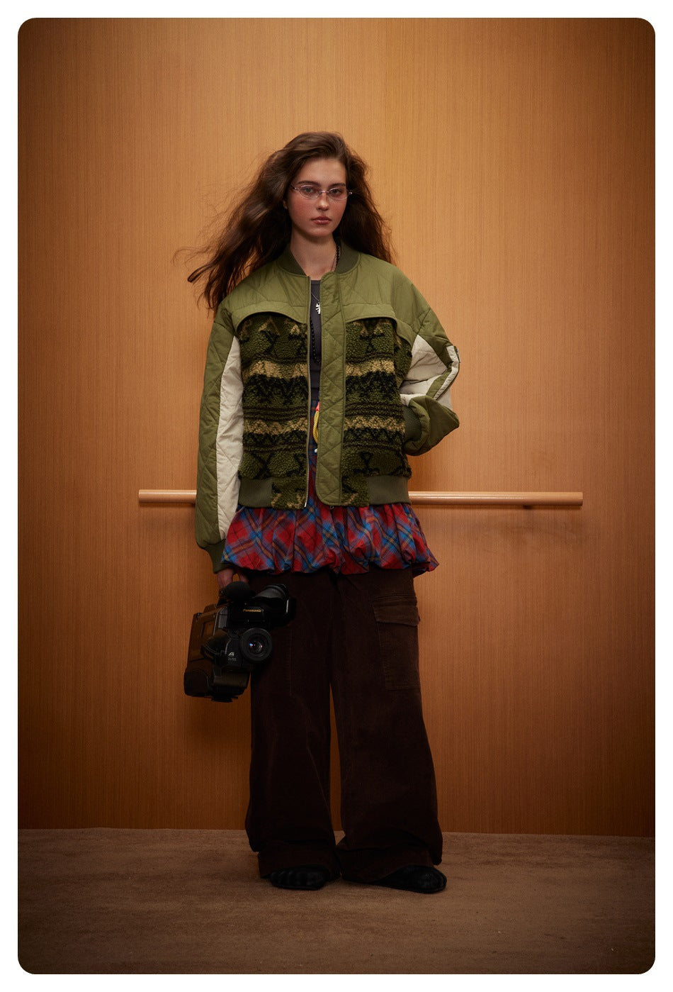 Model wearing the olive quilted bomber jacket with patterned fleece-like panel over a plaid skirt and brown wide-leg pants