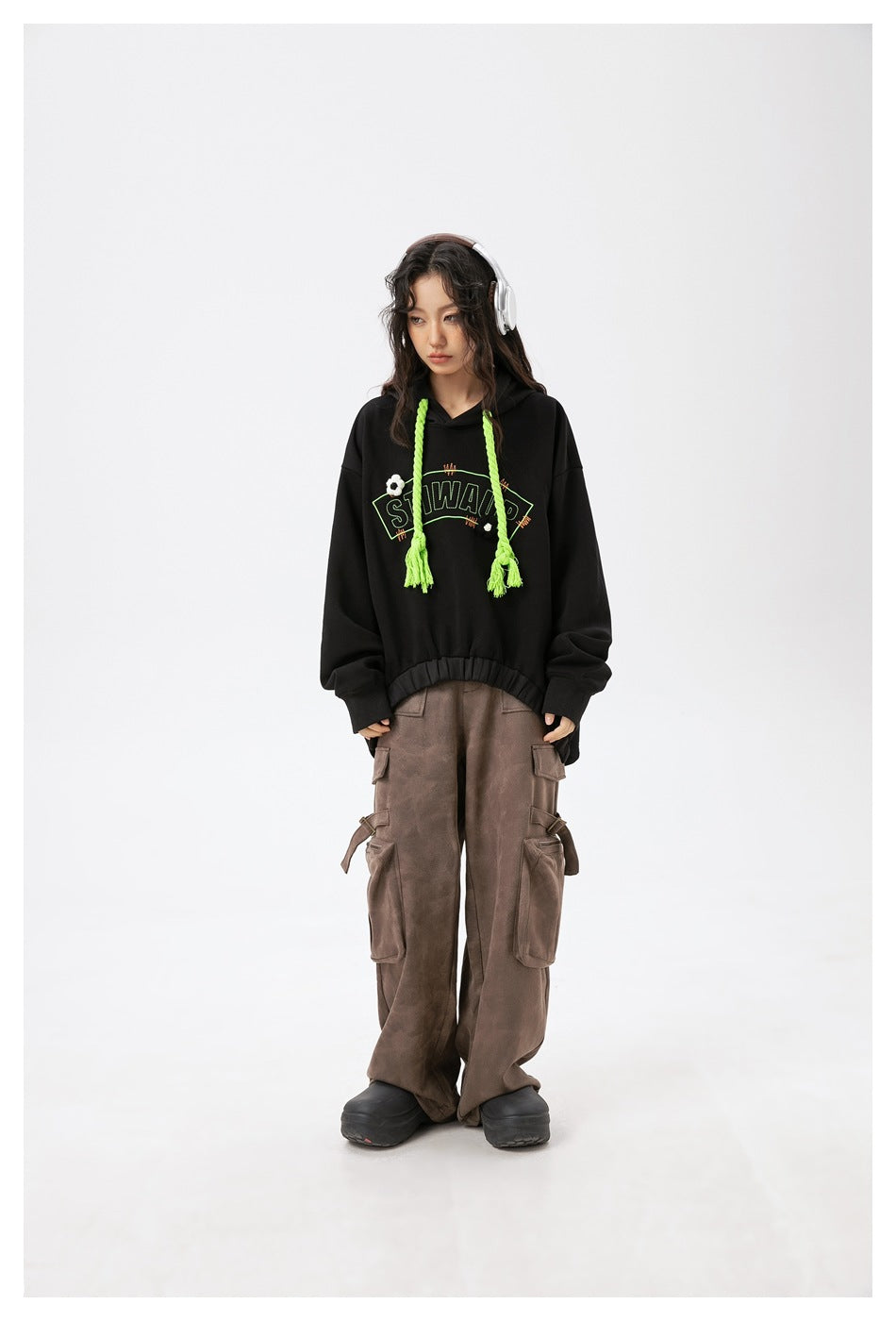 Model wearing an oversized black hoodie with neon green braided drawstrings and graphic, paired with brown cargo pants.