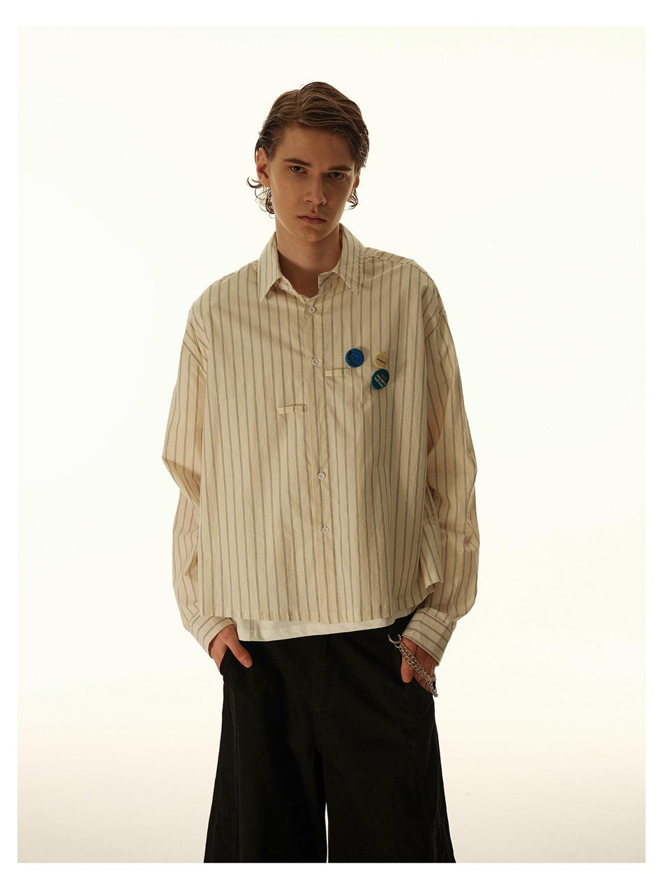 Male model in loose striped cream shirt with badge pins on the chest, hands in pockets of black pants
