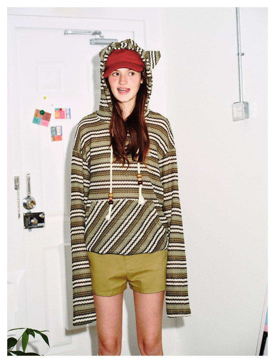 Model wearing the green striped cat-ear hoodie with a red cap and olive shorts, standing against a white door