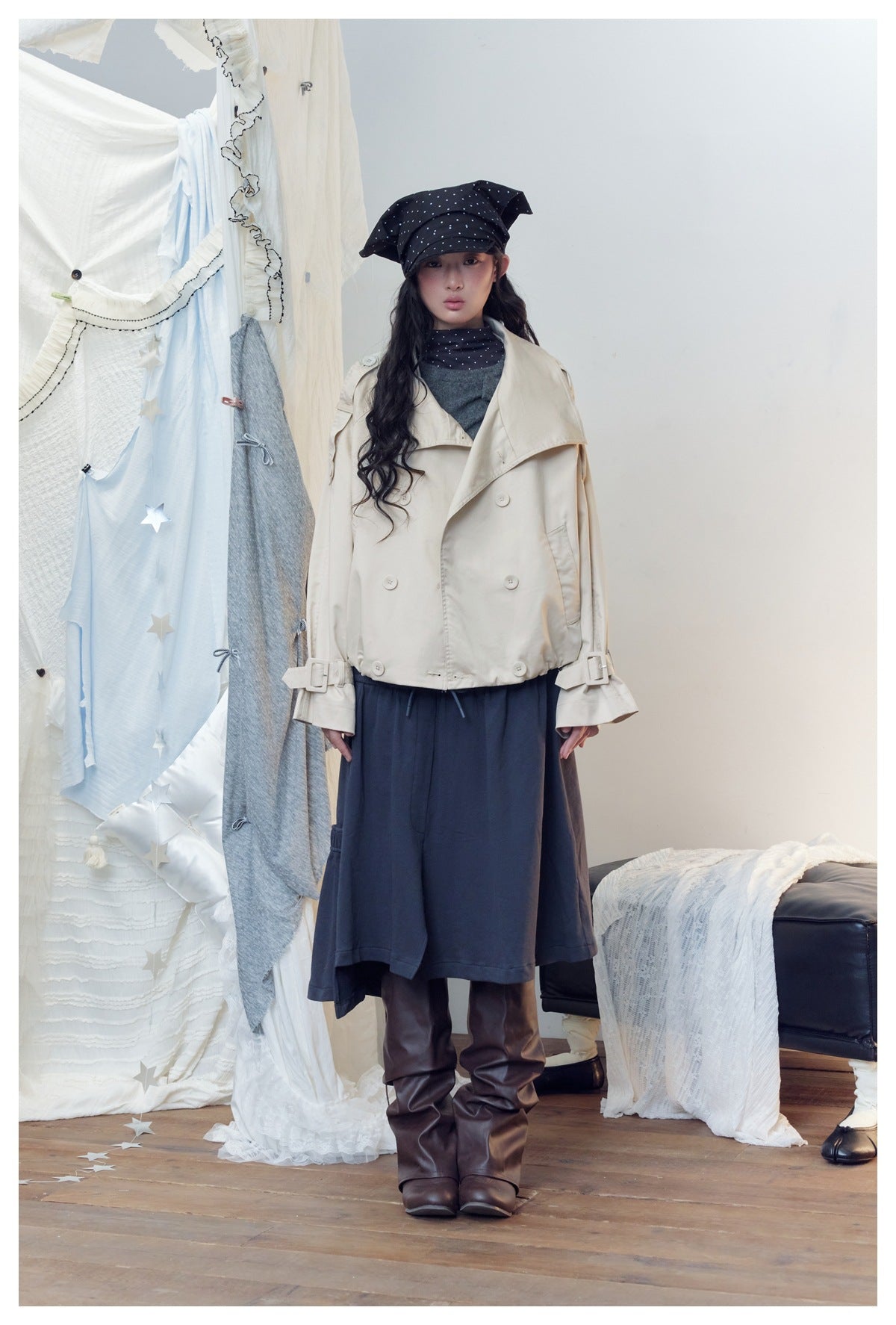 Model wearing a cream short double-breasted trench jacket over a gray top with a navy skirt and brown boots