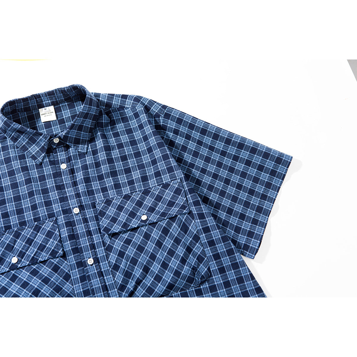 Angled close-up of the navy plaid shirt, showing its short sleeve, button front, and diagonal-plaid pocket flap.
