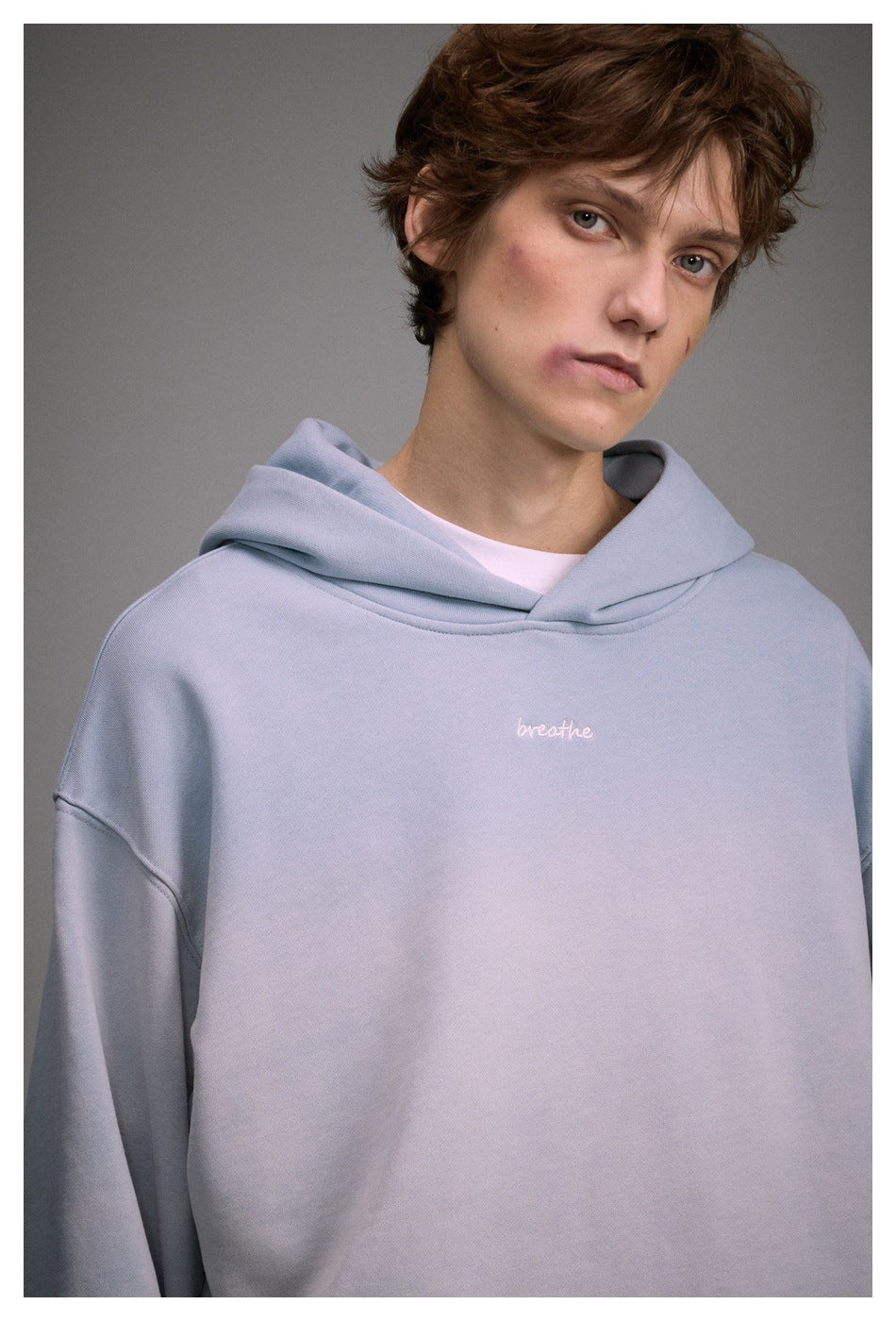 Close-up of a model wearing a pale blue hoodie with a white “breathe” chest embroidery and layered hood.