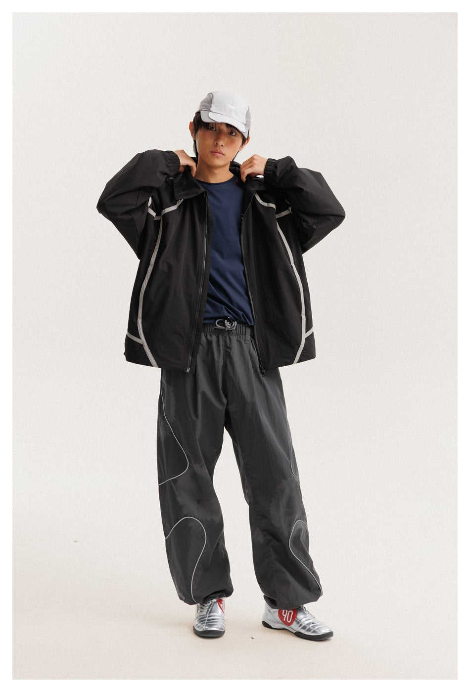 Full-length model wearing an open black hooded jacket with light piping and charcoal pants with curved accents.