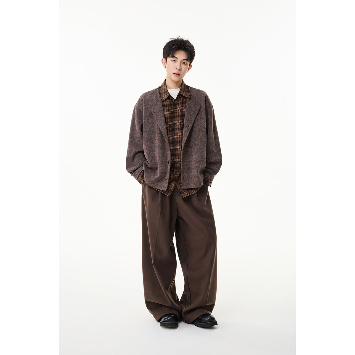 Model wearing a brown-gray button jacket with plaid shirt accents, wide brown trousers and black shoes.