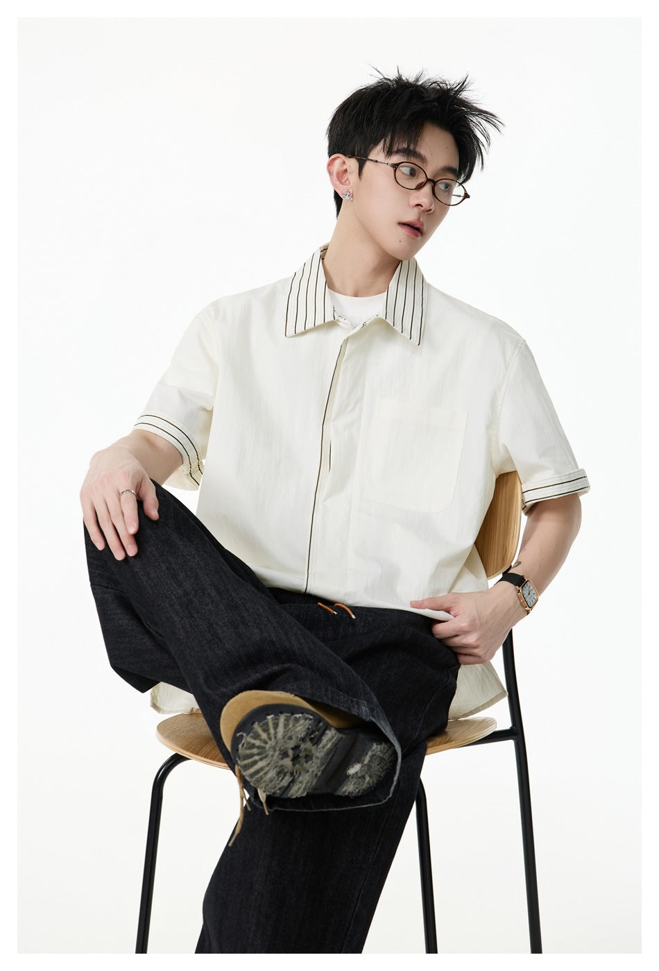 Man seated on a chair wearing an off-white short-sleeve shirt with striped collar and cuffs, black jeans and boots
