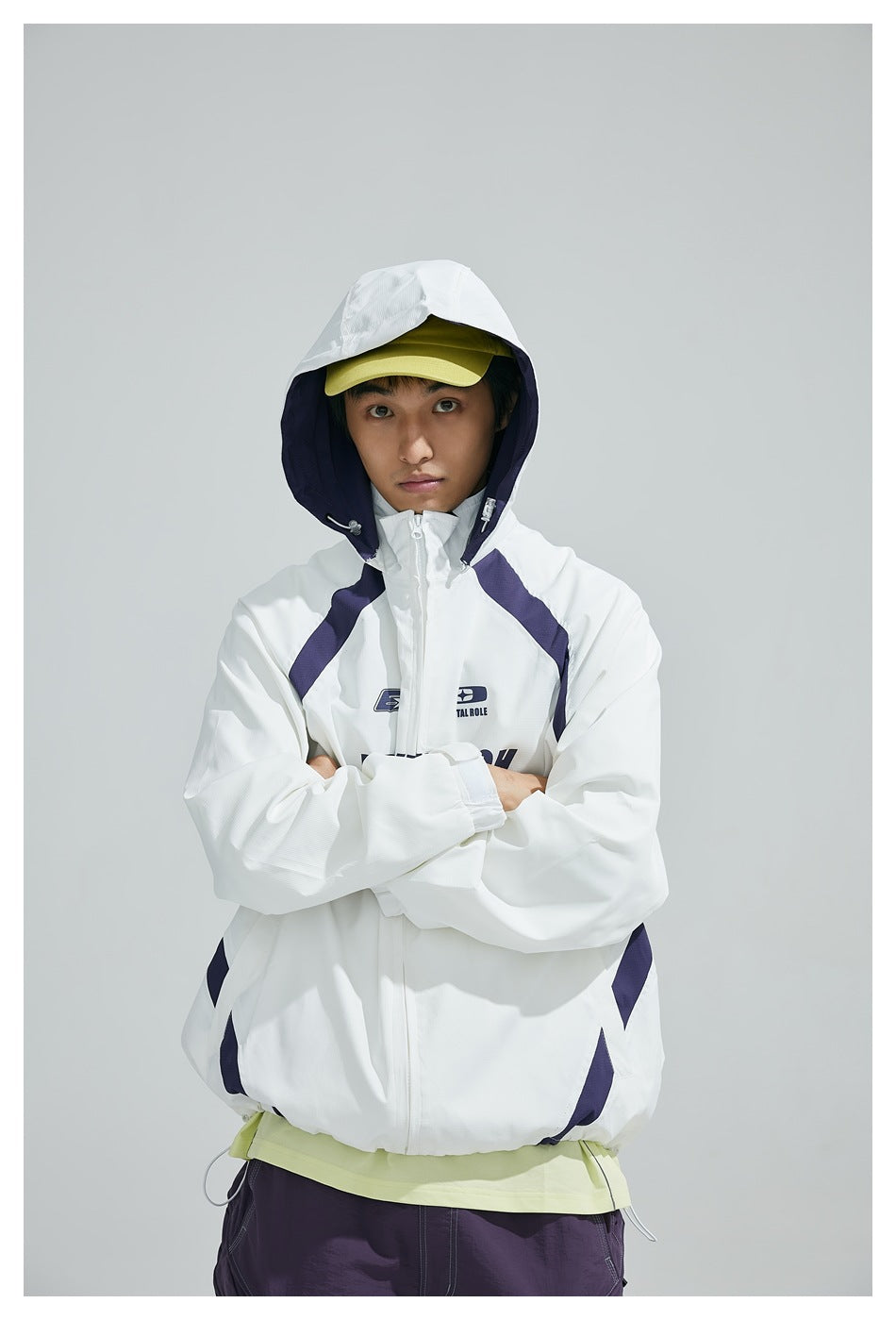 Model wearing a white zip-up hooded jacket with navy panels, arms crossed; a yellow cap and purple trousers are also visible.
