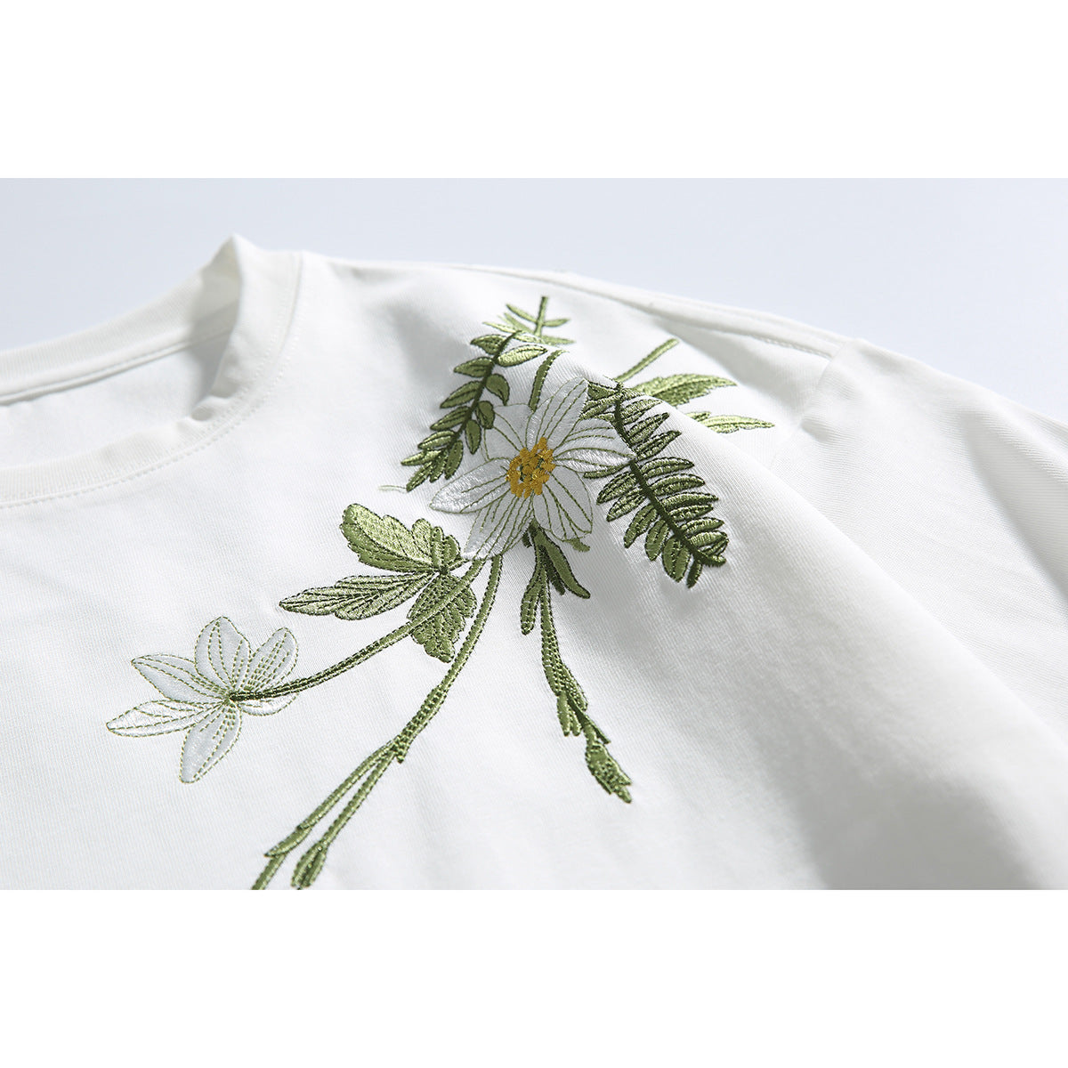 Close-up of a white T-shirt with embroidered white flowers and green leaves across the shoulder and chest.